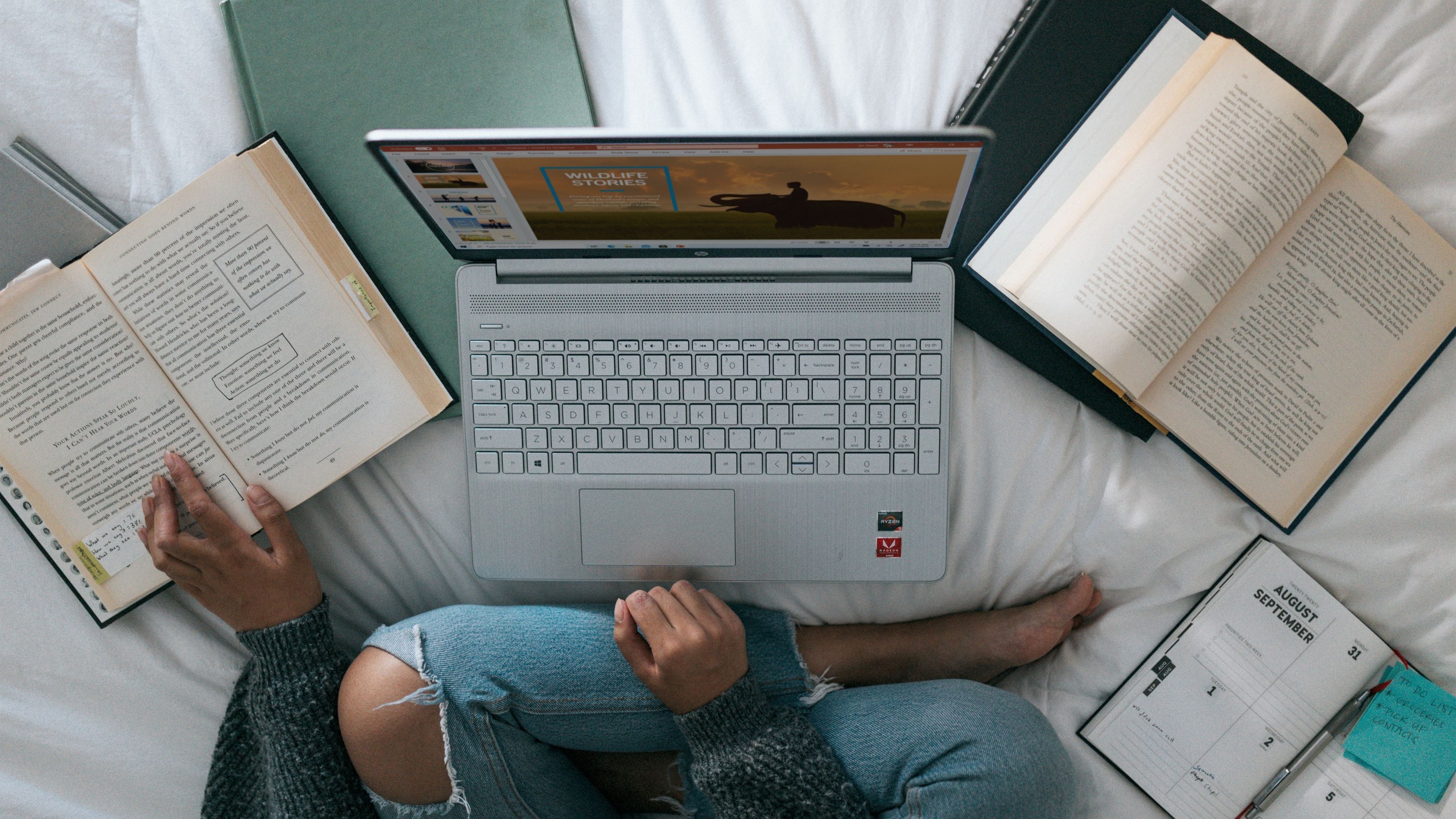 Estudiante usando un portátil rodeada de libros en una cama