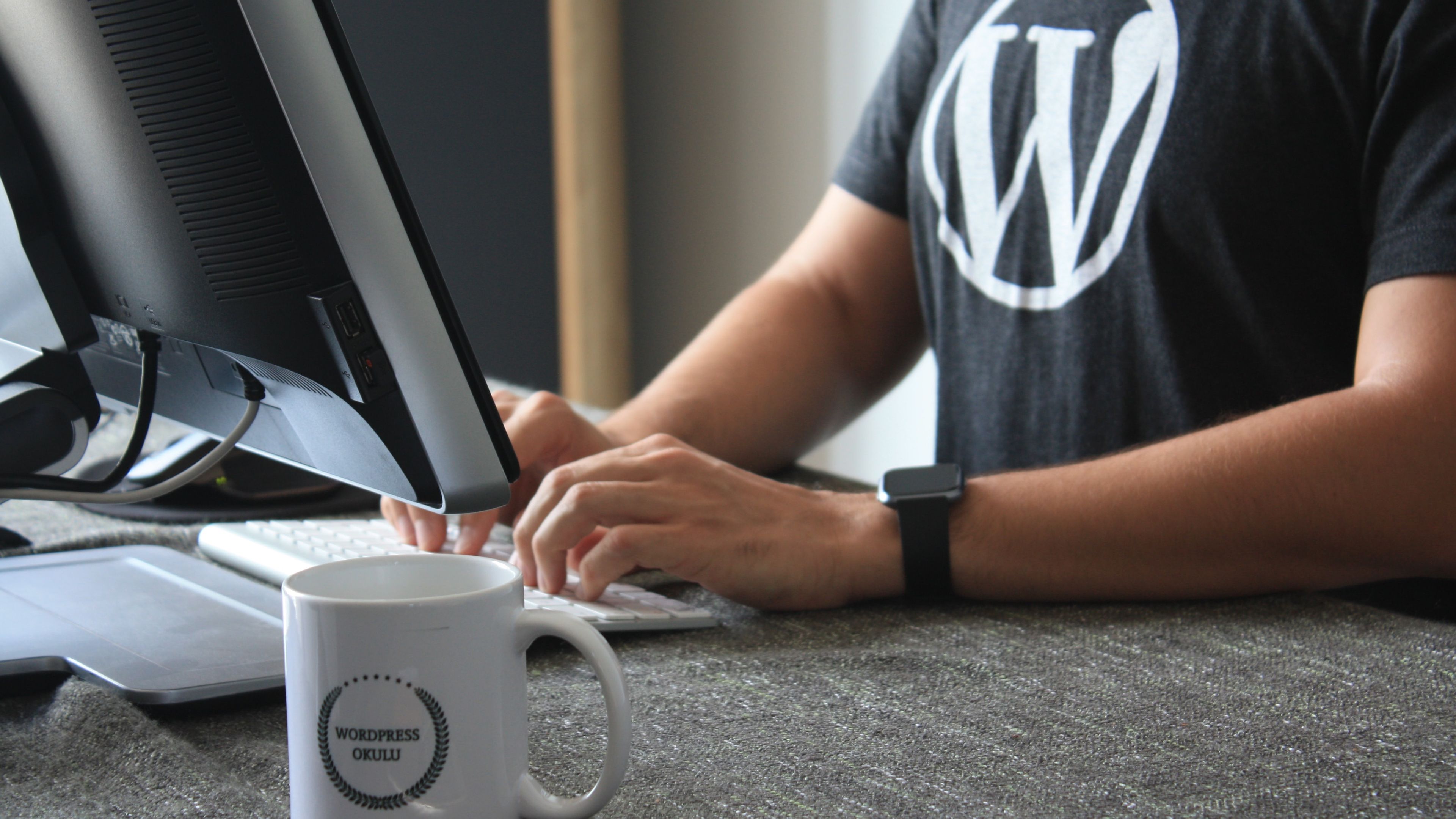 Persona escribiendo en un ordenador con una camiseta con el logo de WordPress y una taza en primer plano